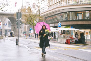 Lozan İsviçre 'nin yağmurlu sokaklarında pembe şemsiyeli ve çiçeklerle yürüyen genç Latin bir kadın, Avrupa' nın nemli havasında şehir yaşamından zevk alan şık bir kadın.