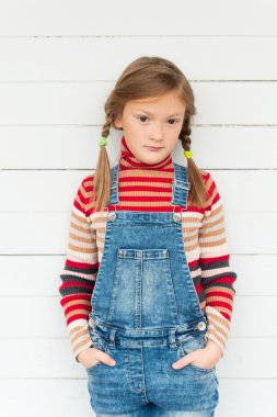 Moda portre şerit rulo boyun kazak ve denim tulum giyen sevimli küçük bir kızın beyaz arka plan karşı