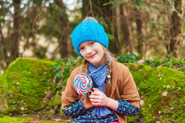 Outdoor portrait of a cute little girl of 7 years old, playing in the park, wearing warm knitted blue hat, holding colorful candy — Stock Photo, Image