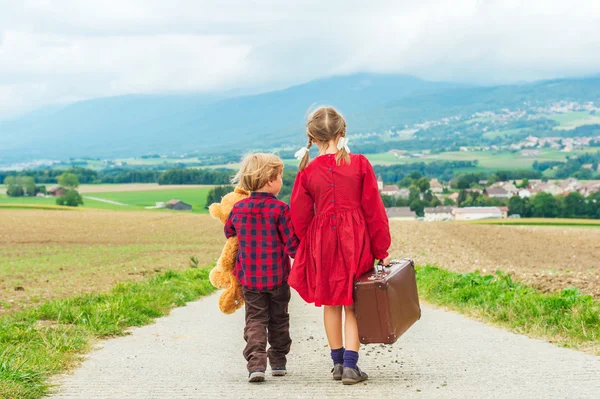 Two cute kids, little girl and her brother, walking down the small road, holding big teddy bear and old brown suitcase