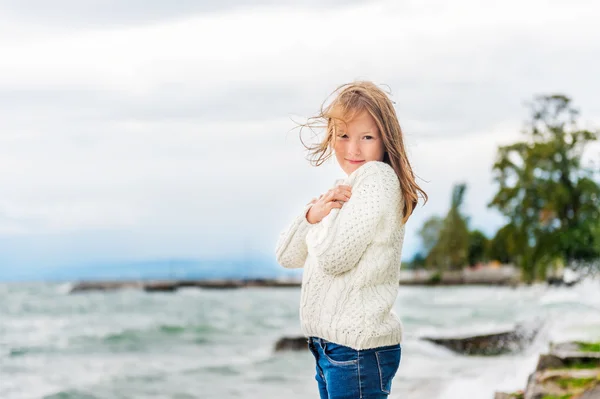 Niedliches kleines Mädchen von 8 Jahren, das an einem sehr windigen Tag am See spielt und einen warmen weißen Strickpullover trägt — Stockfoto