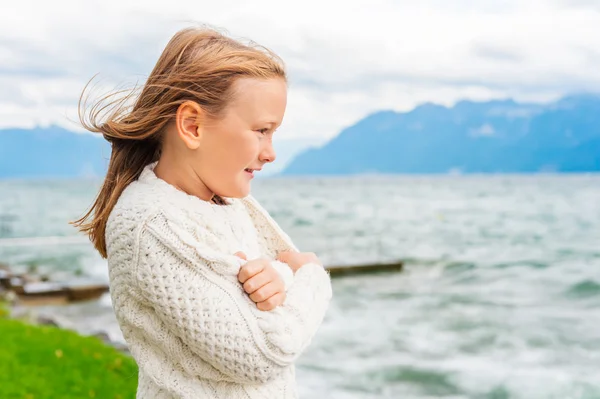 Menina bonito de 8 anos de idade jogando junto ao lago em um dia muito ventoso, vestindo pulôver branco quente de malha — Fotografia de Stock