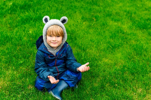 Cute kid resting outdoors, sitting on a lawn, playing on a rainy day, wearing blue waterproof all-in-one suit — Stockfoto