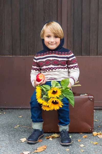 Ritratto autunnale di adorabile bambino biondo di 4 anni, che indossa pullover caldo con cappuccio, jeans in denim scuro e scarpe blu, con una mela e bouquet di girasoli gialli — Foto Stock