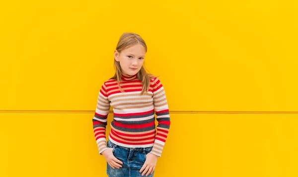 Ritratto di una graziosa bambina di 8 anni, che indossa pullover girocollo a righe e gonna in denim, in piedi accanto alla parete gialla brillante — Foto Stock