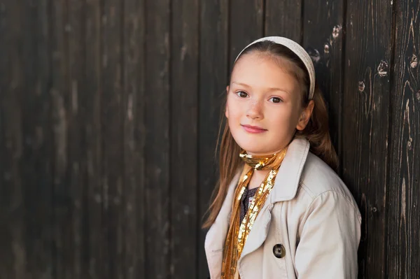トレンチ コートと暗い木製の壁の横に立っている金色のスカーフを身に着けているかわいい女の子の屋外のポートレート — ストック写真