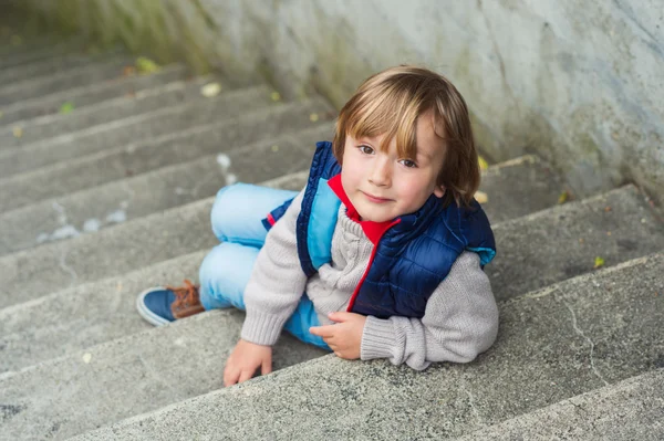 Retrato de moda de um menino loiro bonito sentado em escadas em uma cidade, vestindo pulôver bege e casaco azul — Fotografia de Stock