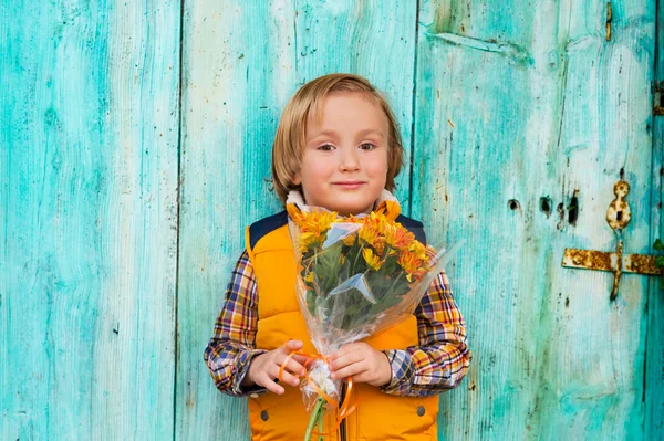 Retrato de outono de adorável menino loiro de 4 anos de idade, vestindo casaco de colete amarelo quente, segurando pequeno buquê de flores de crisântemo laranja, em pé na frente da parede de madeira turquesa — Fotografia de Stock