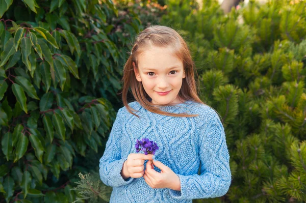 Ritratto all'aperto di una graziosa bambina di 7 anni, vestita di pullover blu, che tiene in mano le prime viola dei fiori primaverili — Foto Stock