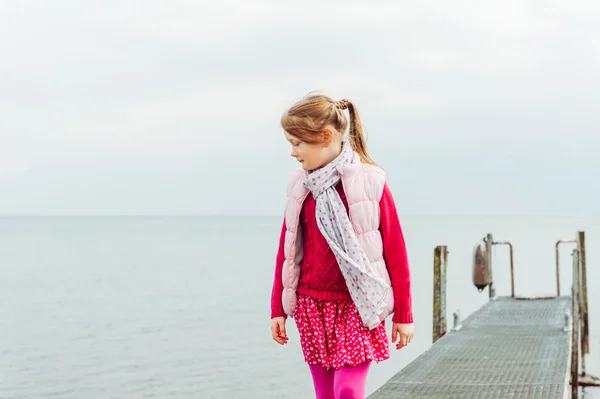 Außenporträt eines niedlichen kleinen Mädchens, das an einem bewölkten Tag am See spielt, rotes Kleid und rosa Weste trägt, getöntes Bild — Stockfoto