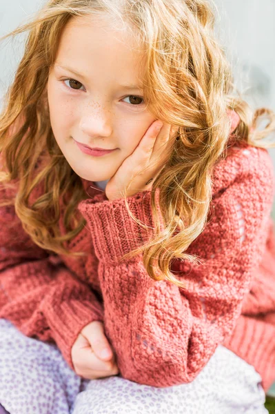 Retrato de cerca de una linda niña de 8 años con el pelo rizado, usando jersey de terracota, sentado sobre fondo de madera gris —  Fotos de Stock