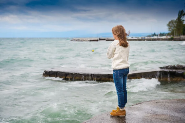 Niedliches kleines Mädchen von 8 Jahren, das an einem sehr windigen Tag am See spielt; es trägt einen warmen weißen Strickpullover; Jeans und Stiefel — Stockfoto