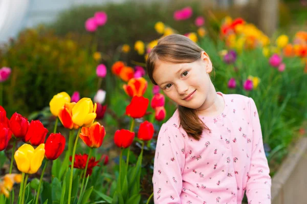 Outdoor portrait of a cute little girl of 7 years old, wearing pink jacket, sitting next to tulips — Stock Photo, Image