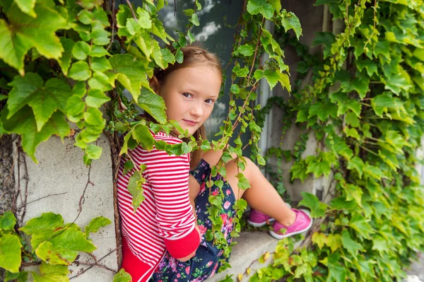 Outdoor Portret van een schattige jonge meisje zittend op de vensterbank met ivy, dragen kleurrijke rok, rode en witte streep vest en roze laceless schoenen — Stockfoto