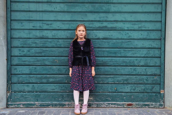 Ritratto di moda all'aperto di una bambina carina di 8 anni, vestita di nero e pelliccia sintetica, in piedi accanto al muro di legno verde — Foto Stock