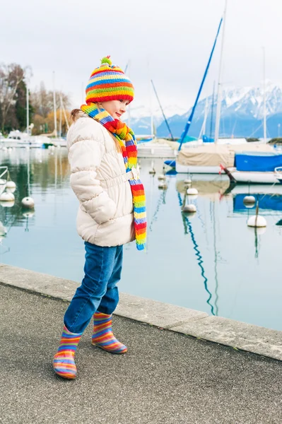 Adorable little girl plying by the lake in early spring, wearing white warm jacket, colorful set of scarf and hat and rain boots, vertical image — Stok fotoğraf