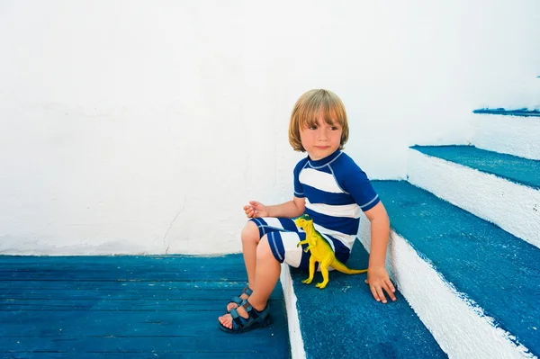 Adorable niño jugando con un juguete de dinosaurio, usando divertido traje de baño y zapatos de playa, sentado en escaleras azules, concepto de vacaciones de verano —  Fotos de Stock