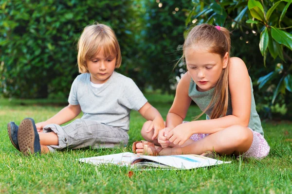 Two adorable kids, little boy and his sister reading a book in garden on a nice warm sunny day — Stock Photo, Image