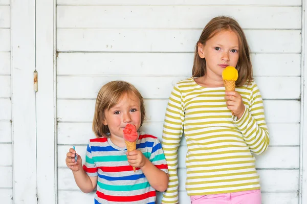 Retrato de duas crianças adoráveis comendo sorvete colorido ao ar livre, de pé ao lado de fundo de madeira branca — Fotografia de Stock