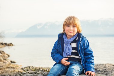 Outdoor portrait of adorable little boy of 4-5 years old, resting by the lake, wearing warm blue jacket and scarf