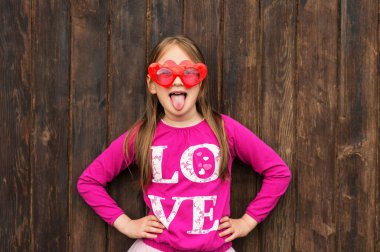 Funny portrait of a cute little girl wearing big heart shaped sunglasses, pulling tongue