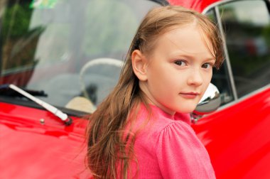 Outdoor close up portrait of a cute little girl wearing bright pink pullover, standing next to red car