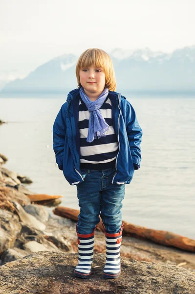 Outdoor portrait of adorable little blond boy of 4-5 years old, having fun by the lake on a nice sunny spring day, wearing warm blue jacket, scarf, denim jeans and stripes rain boots — Stock fotografie