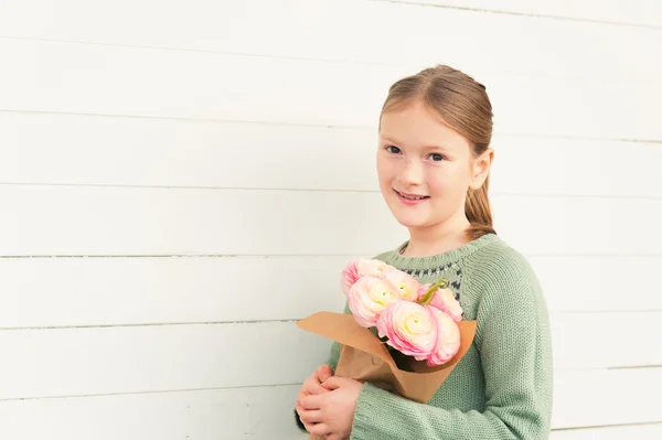 Porträtt av bedårande liten flicka på 8-9 år gammal, bär varma gröna pullover, håller våren rosa blommor, stående mot vit trä bakgrund, mors dag koncept — Stockfoto