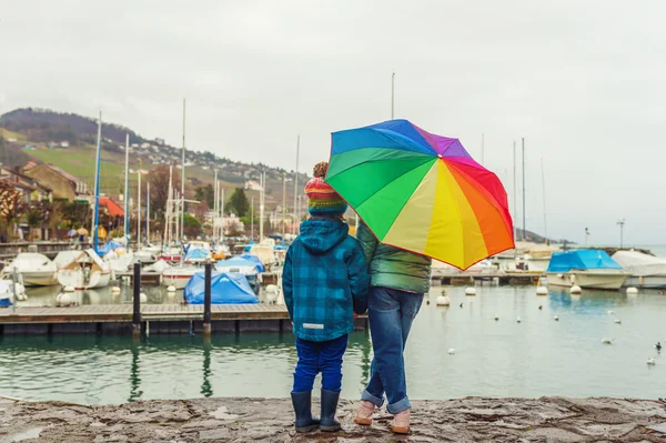 Открытый портрет двух милых детей, отдыхающих у озера в дождливый день, прячущихся под большим красочным зонтиком — стоковое фото