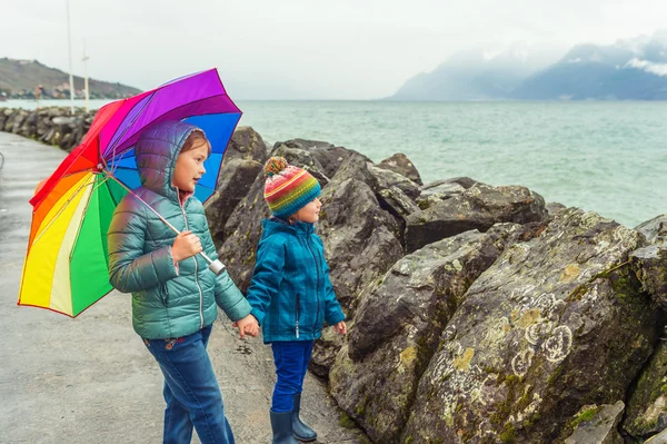 Outdoor portrait of two cute kids resting by the lake on a rainy day, hiding under big colorful umbrella — Stock Photo, Image
