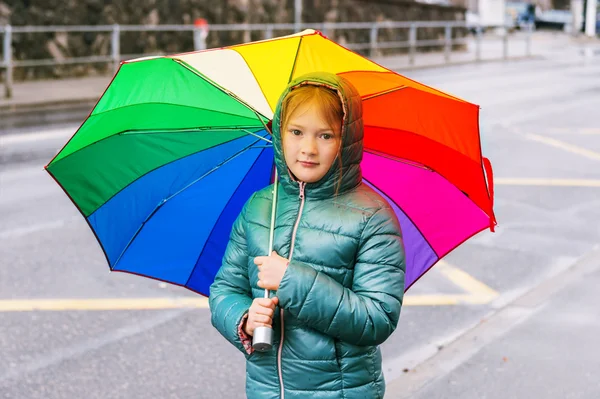 Outdoor portrait of a cute little girl, wearing warm green jacket, holding big colorful umbrella — Stock Photo, Image