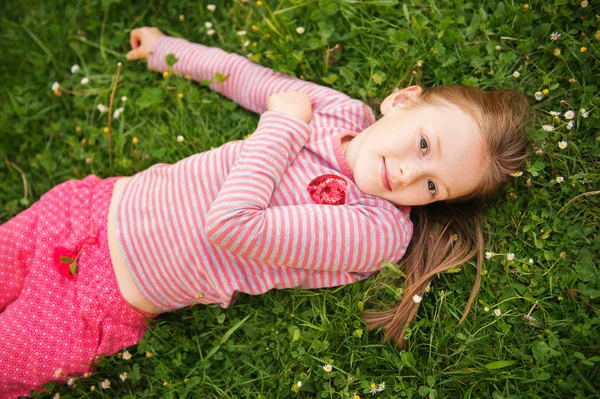 Portrait of a cute little girl of 7 years old in the park, laying on grass, wearing red clothes — ストック写真