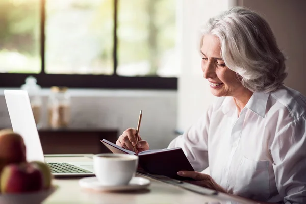 Señora de pelo gris tomando notas con lápiz y utilizando el ordenador portátil — Foto de Stock