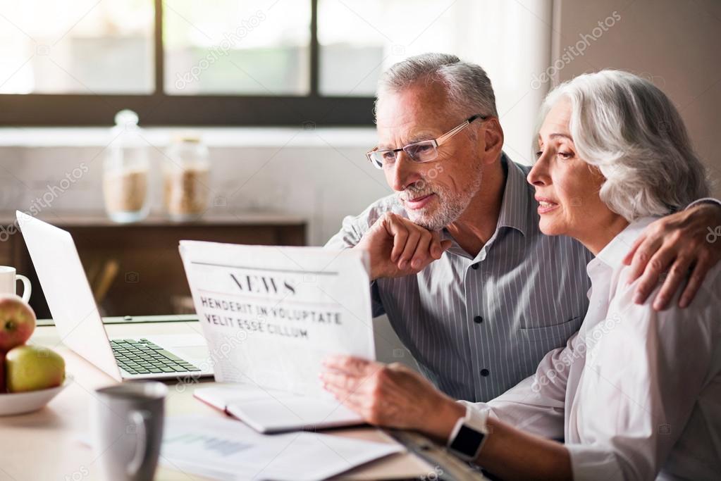 Happy and caring old couple reading newspaper at the kitchen