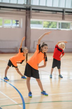 Spor salonunda egzersiz yapan ve sola doğru eğilen üç çocuk.