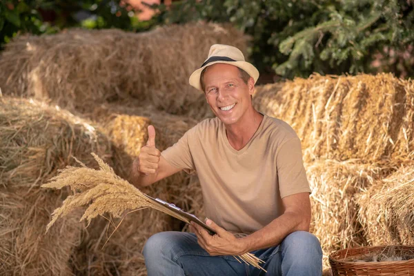 Hombre agricultor sosteniendo la carpeta de clip y manojo de espiguillas de mijo, mostrando el pulgar hacia arriba — Foto de Stock