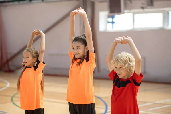 Three kids in bright tshirts doing stretching in the gym — Stock Photo, Image