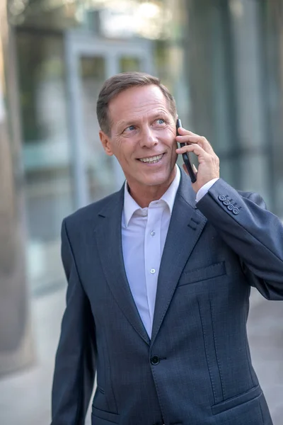 Uśmiechnięty dojrzały biznesmen wychodzący na zewnątrz, rozmawiający przez telefon — Zdjęcie stockowe