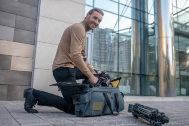 Gülümseyen kameraman dışarıda dizinin üzerinde duruyor, ekipmanları çıkarıyor.