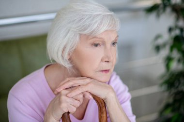 Pembe tişörtlü gri saçlı kadın ciddi ve düşünceli görünüyor.