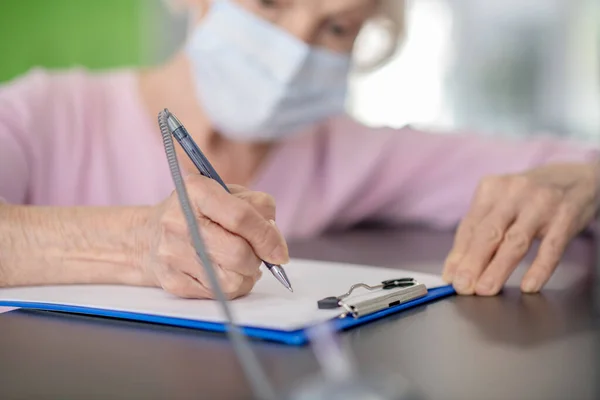 Grey-haired womanin facial mask signing the health insurance contract — Stock Photo, Image