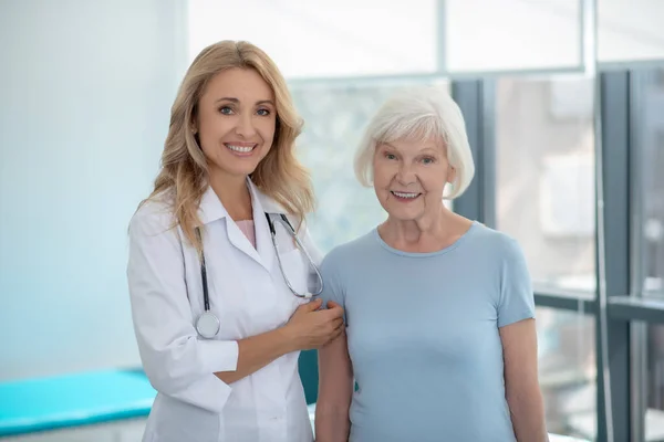 Длинноволосая милая улыбающаяся медсестра, стоящая рядом со своим пожилым пациентом — стоковое фото