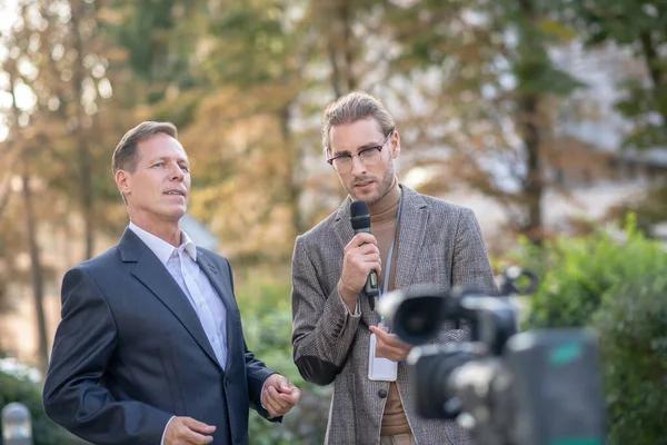 Fair-haired male journalist interviewing mature male in front of camera