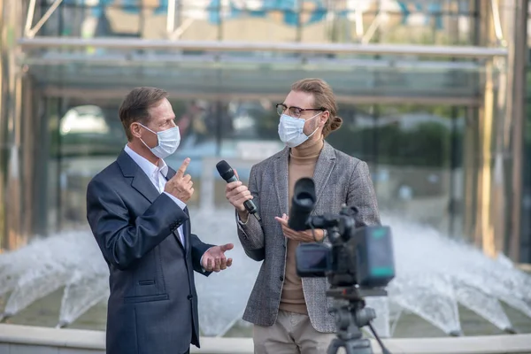 Fair-haired male journalist interviewing mature male in front of camera, both wearing masks