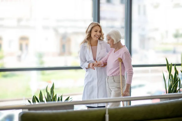 Doctora y una anciana con una muleta caminando por un pasillo y conversando — Foto de Stock