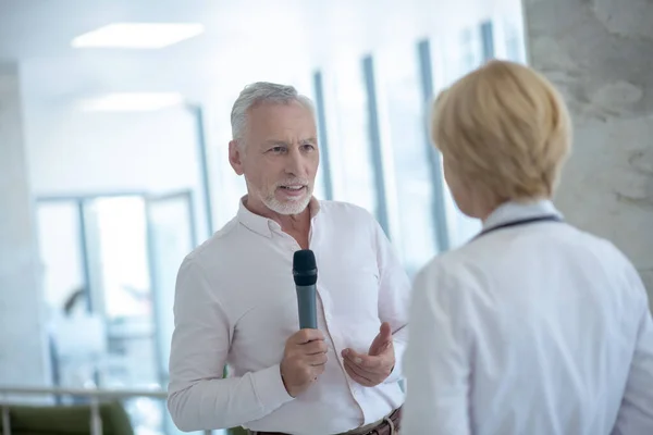 Periodista masculino de cabello gris sosteniendo micrófono, entrevistando a doctora rubia — Foto de Stock