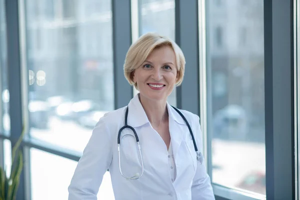 Smiling blonde female doctor standing next to the window