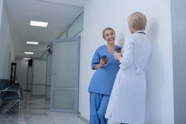 Två leende kvinnliga läkare lutar sig mot väggen, dricker kaffe, diskuterar — Stockfoto