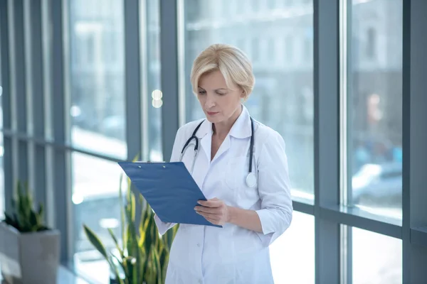 Blonde female doctor standing next to the window, taking notes