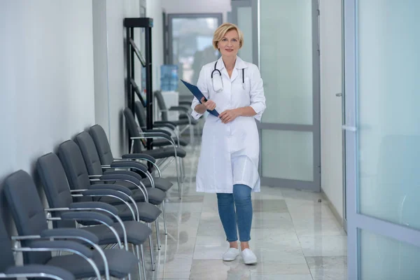 Улыбающаяся блондинка-врач со стетоскопом идет по коридору, держа папку с клипом — стоковое фото